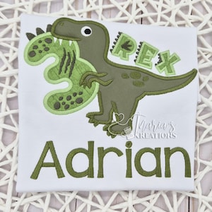 May include: A white t-shirt featuring an embroidered green dinosaur design. The dinosaur is biting a large green number three. The text REX is embroidered above the dinosaur's tail, and the name Adrian is embroidered in green below the dinosaur.