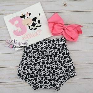 May include: A white t-shirt with a pink number 3 and a cow graphic. The shirt is personalized with the name "Mia". A pair of black and white cow print bloomers and a pink bow are also pictured.