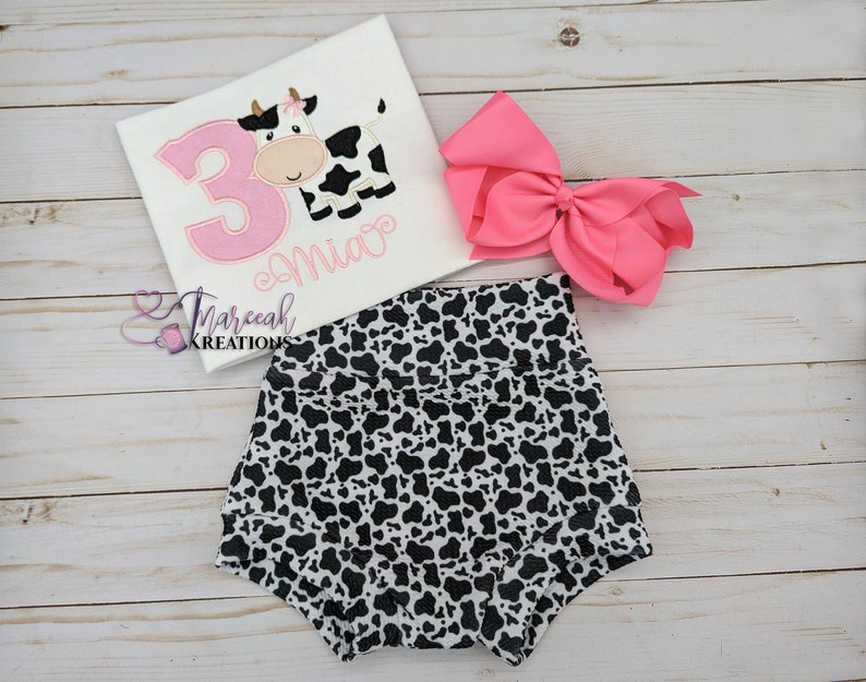 May include: A white t-shirt with a pink number 3 and a cow graphic. The shirt is personalized with the name "Mia". A pair of black and white cow print bloomers and a pink bow are also pictured.