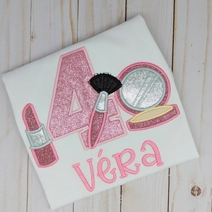 May include: Pink glitter number four with a pink lipstick, a pink makeup compact with a silver mirror, and a pink makeup brush. The name "Vera" is written in pink below the number.