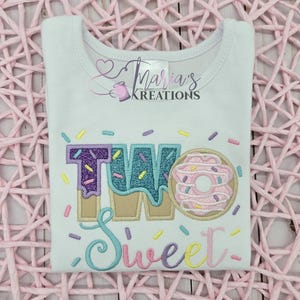 May include: A white short-sleeved t-shirt featuring an embroidered design with the text TWO Sweet. The word TWO is rendered in glittery purple and blue fabric with a donut graphic, while Sweet is in cursive. Colorful sprinkles surround the text.