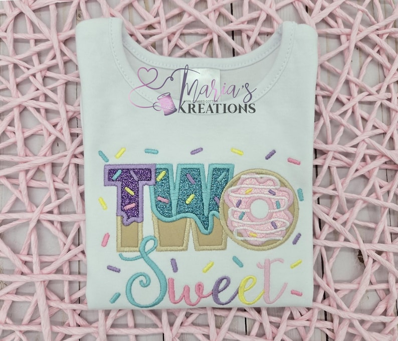May include: A white short-sleeved t-shirt featuring an embroidered design with the text TWO Sweet. The word TWO is rendered in glittery purple and blue fabric with a donut graphic, while Sweet is in cursive. Colorful sprinkles surround the text.