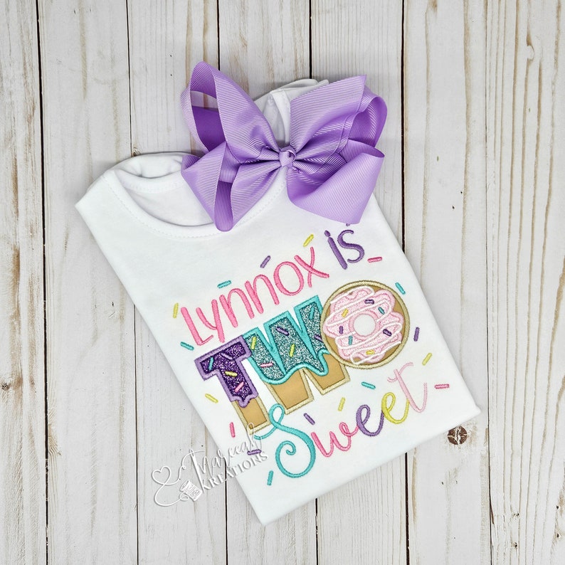 May include: A white short-sleeved shirt featuring embroidered text that reads "Lynnox is TWO Sweet" with a donut graphic. The shirt is decorated with colorful sprinkles and includes a large, solid light purple ribbon hair bow attached at the top.