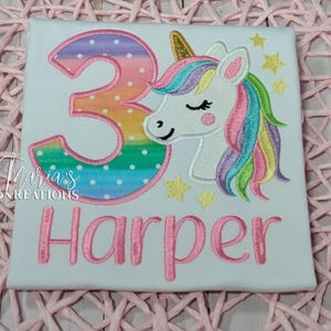 May include: A white t-shirt featuring an embroidered design of a large number 3 with a rainbow gradient and white polka dots, next to a white unicorn with a gold horn and a multi-colored mane. The name Harper is embroidered in pink below.