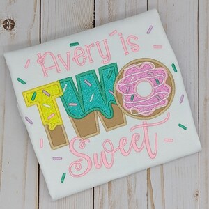 Może przedstawiać: Biała koszulka z haftem w kolorze r&oacute;żowym, zielonym i ż&oacute;łtym, na kt&oacute;rej widnieje napis "Avery is Two Sweet" oraz grafika przedstawiająca pączka.