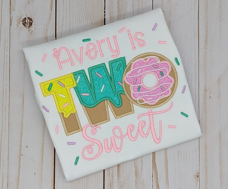 Może przedstawiać: Biała koszulka z haftem w kolorze r&oacute;żowym, zielonym i ż&oacute;łtym, na kt&oacute;rej widnieje napis "Avery is Two Sweet" oraz grafika przedstawiająca pączka.