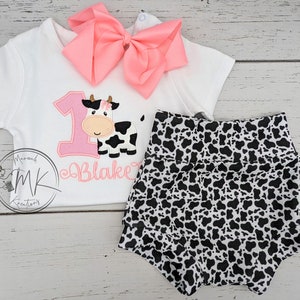 May include: A white onesie with a pink cow and the number one embroidered on it. The onesie also has the name "Blake" embroidered on it. The onesie is paired with black and white cow print shorts.