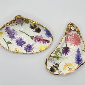 May include: Two decorative oyster shell trinket dishes. The shells are painted white with floral and bee designs in purple, pink, and yellow. The edges are trimmed with gold paint.