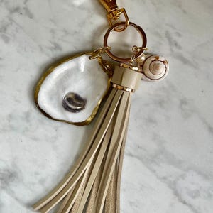May include: A keychain featuring a gold-tone lobster clasp and ring, a white oyster shell with a gold-painted edge and a dark center, a beige leather tassel, and a small spiral seashell charm. The hardware is metallic gold, and the tassel is long.