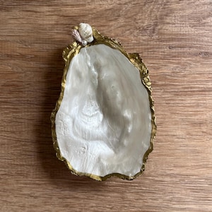 May include: A decorative oyster shell dish with a gold-painted rim and a pearlescent white interior. Small seashells and a pearl accent the top edge. The dish is displayed on a wooden surface.