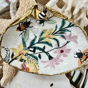 May include: A decorative seashell with a gold-painted rim and a fabric interior featuring a floral and bee design. The shell is adorned with a jeweled bee and is set against a backdrop of coral and other shells.