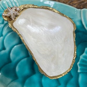 May include: A decorative oyster shell dish featuring a smooth, pearlescent white interior and a metallic gold-painted rim. The shell is adorned with a small, textured starfish embellishment covered in tiny white beads and two small pearls.