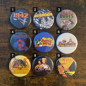 May include: Nine colorful round buttons featuring classic arcade game designs. The buttons display game titles like "1942", "Asteroids", "Bad Dudes", "Berzerk", "Bubble Bobble", "Burger Time", "Centipede", "Contra", and "Defender".