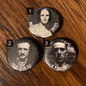 May include: Three black and white button pins featuring portraits of Mary Wollstonecraft Shelley, Edgar Allan Poe, and H.P. Lovecraft. The pins are on a wooden surface.