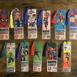 May include: A collection of Marvel Comics Group bookmarks featuring various superheroes, including Spider-Man, Thor, and Iron Man. Each bookmark has a tassel and comic book cover art. The bookmarks are on a wooden surface.