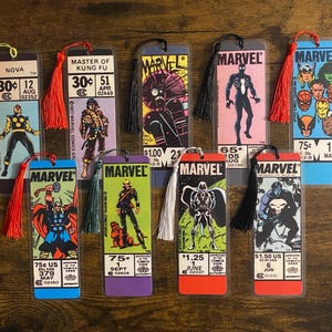 May include: A collection of ten rectangular Marvel comic book bookmarks, each encased in clear plastic and featuring a colorful tassel. The designs display various comic book covers, character illustrations, price tags, and dates from the Marvel series.