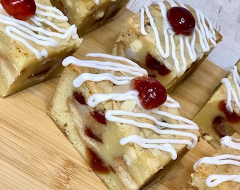 May include: Close-up of several square-shaped baked goods on a wooden cutting board. The treats have a light-colored base with visible cherries, almond slices, and white icing drizzle. Each is topped with a bright red cherry.