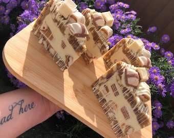 May include: Four slices of a layered dessert on a wooden serving board. The dessert features a creamy white filling, chocolate pieces, and white chocolate drizzle. The background includes purple flowers.