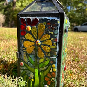 Puede incluir: Linterna de metal negro con un diseño floral de mosaico. El mosaico presenta piezas de vidrio amarillas, rojas y verdes dispuestas en un patrón de flores. La linterna tiene una puerta con bisagras y una parte superior de vidrio, adecuada para la decoración exterior.