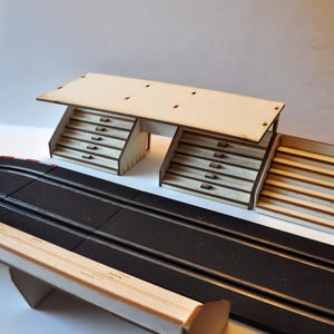 Può includere: Un modello in legno di una tribuna con tre sezioni, ciascuna con posti a sedere a gradini. La tribuna è in legno marrone chiaro e ha un tetto piatto. Il modello è posizionato su una pista nera con linee bianche.