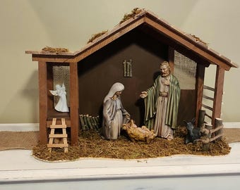 May include: A detailed Nativity scene featuring a wooden stable with a moss-covered roof. Inside, figurines depict Mary, Joseph, and baby Jesus in a manger. An angel, a small ladder, and a dog are also present.