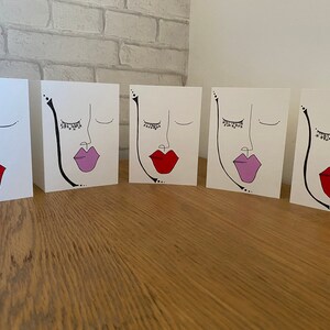 May include: Five blank greeting cards featuring a simple line drawing of a face with closed eyes and eyelashes. Each card has a different coloured lip, red, pink, and purple.