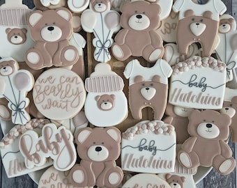 May include: A selection of baby-themed biscuits, featuring teddy bears, baby bottles, and babygrows. Decorated with white and brown icing, some biscuits have the words "Baby," "Baby Hutchins," and "We can bearly wait." A delightful treat for a baby shower or new arrival.
