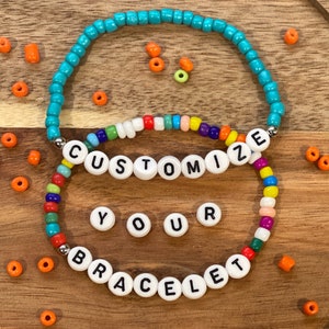 May include: Two colorful beaded bracelets with white letter beads spelling "CUSTOMIZE YOUR BRACELET". The bracelets are on a wooden surface with loose beads scattered around.