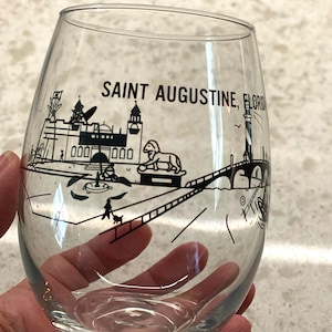 May include: A clear glass wine glass with a black and white illustration of the St. Augustine, Florida skyline. The illustration includes a lighthouse, a bridge, a lion statue, and a building with the text "000000" on it. The text "SAINT AUGUSTINE, FLORIDA" is printed on the glass.