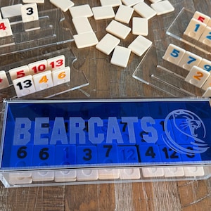 May include: A Rummikub game set featuring off-white numbered tiles, clear acrylic tile racks, and a blue acrylic storage box with the word BEARCATS and a lion logo etched on the front. The set includes numerous loose tiles scattered on the surface.