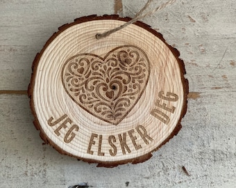 May include: A wooden round ornament with a heart design and the text "JEG ELSKER DEG" engraved on it. The ornament is attached to a string.