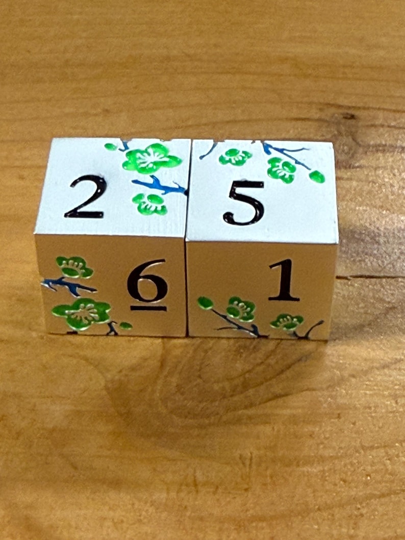 May include: Two white wooden dice with black numbers and green floral designs. The dice show the numbers 2, 5, 6, and 1. The floral designs are green with blue stems.