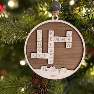 May include: A round wooden Christmas ornament with a white border and a dark wood center. The ornament features Scrabble tiles spelling out names and the text "The Kruger's 2025". It hangs from a twine loop.
