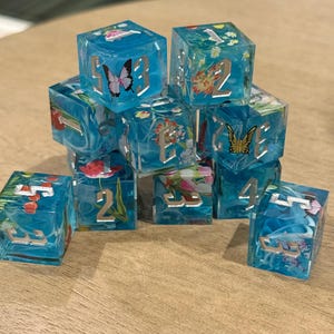 May include: A collection of clear, blue resin dice with embedded floral and butterfly designs. Each die features silver numbering. The dice are arranged on a wooden surface, showcasing their intricate details and vibrant colours.