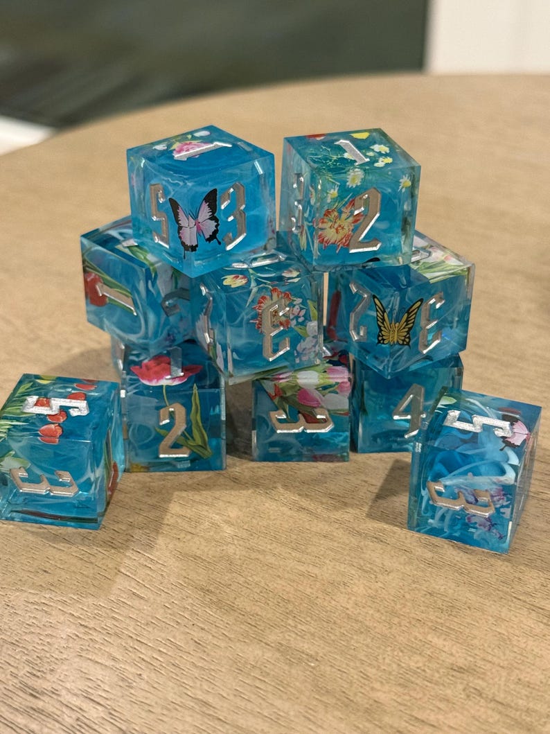 May include: A collection of clear, blue resin dice with embedded floral and butterfly designs. Each die features silver numbering. The dice are arranged on a wooden surface, showcasing their intricate details and vibrant colours.
