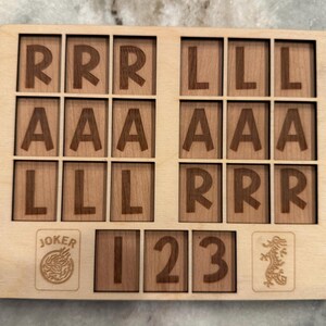 May include: A rectangular light-colored wooden board featuring a grid of eighteen square tiles engraved with the letters R, A, and L. Below the grid are four additional tiles displaying the word JOKER, the numbers 1, 2, and 3, and a dragon icon.