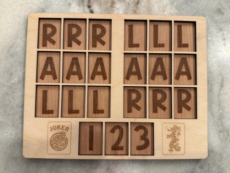 May include: A rectangular light-colored wooden board featuring a grid of eighteen square tiles engraved with the letters R, A, and L. Below the grid are four additional tiles displaying the word JOKER, the numbers 1, 2, and 3, and a dragon icon.