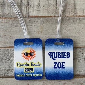 Può includere: Due taglie per bagagli blu e bianche con un effetto sfumato blu. Un'etichetta presenta un sole giallo con occhiali da sole e il testo "Florida Finals 2024 Pinnacle Cheer Memphis". L'altra etichetta presenta il testo "Rubies Zoe" in bianco.