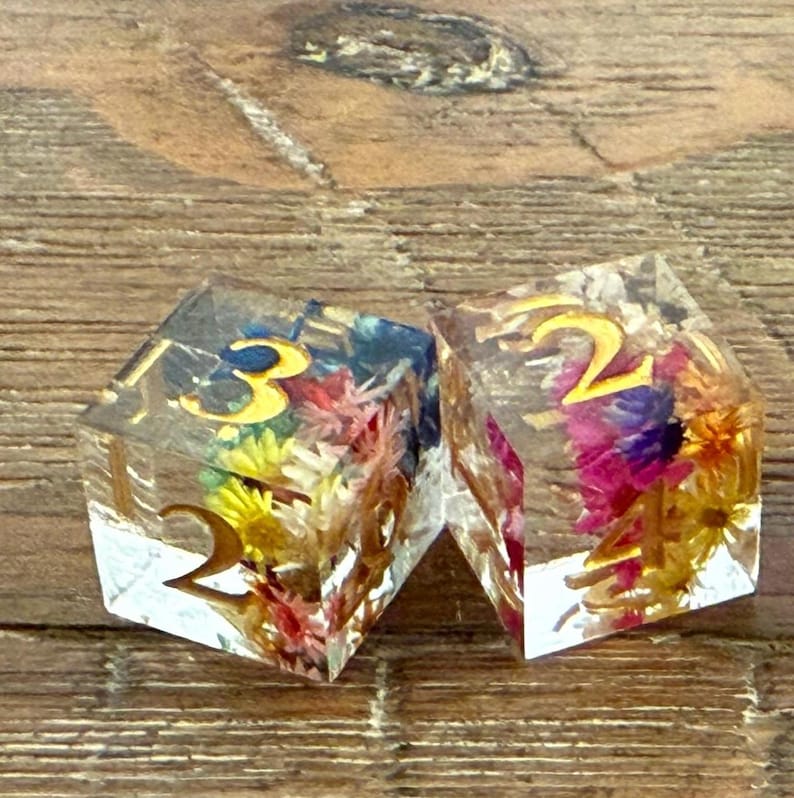 May include: Two clear resin dice with gold numbers and colorful dried flowers inside. The dice are set against a wooden background. The dice are for tabletop games.
