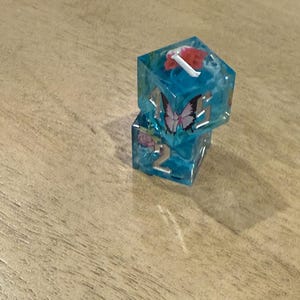 May include: Two stacked, clear resin dice with a blue tint. Each die has embedded floral and butterfly designs. One die has a red flower and white accents. The other die has a pink rose and the number 2.