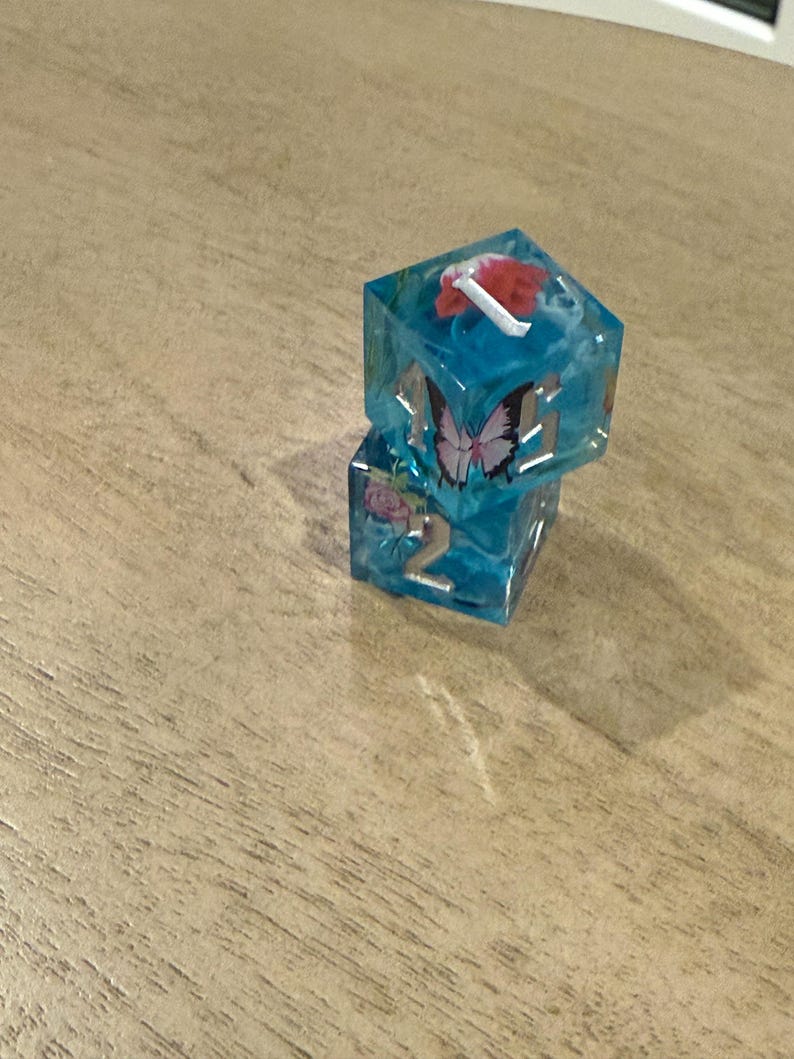 May include: Two stacked, clear resin dice with a blue tint. Each die has embedded floral and butterfly designs. One die has a red flower and white accents. The other die has a pink rose and the number 2.