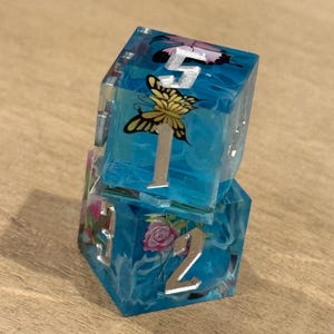 May include: Two stacked, clear resin dice with a blue, swirling interior. Each die face features a silver number and a butterfly or rose design. The dice are on a light brown wooden surface.