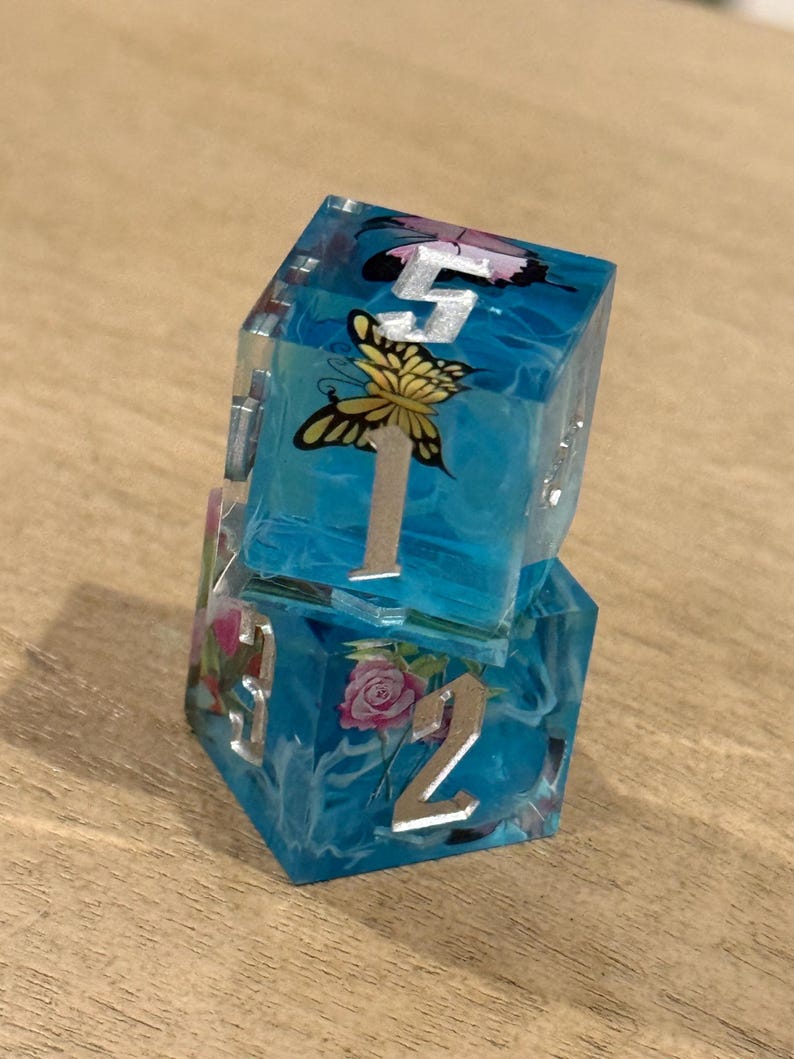 May include: Two stacked, clear resin dice with a blue, swirling interior. Each die face features a silver number and a butterfly or rose design. The dice are on a light brown wooden surface.