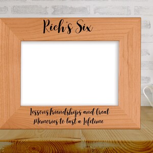 May include: A light brown wooden picture frame with the text "Rich's Six" at the top and "Lessons friendships and great Memories to last a lifetime" at the bottom. The frame is on a wooden surface.
