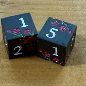 May include: Two black dice with white numbers and red floral accents. The dice show the numbers 1, 2, and 5. The dice are on a wooden surface.