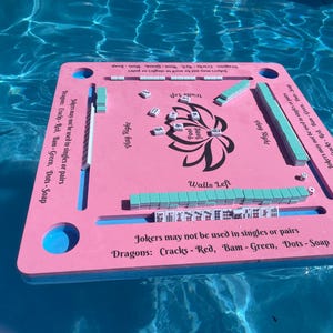 Puede incluir: Tablero de juego flotante rosa y azul con las palabras "Pool Jong" y "Walls Left" impresas en &eacute;l. El tablero tiene un dise&ntilde;o de flor de loto en el centro e instrucciones para jugar al juego.