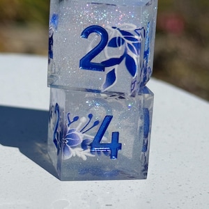 May include: Two clear, square dice stacked on top of each other. The dice have blue numbers and floral designs. The interior of the dice is filled with glitter. The top die shows the number 2, and the bottom die shows the number 4.