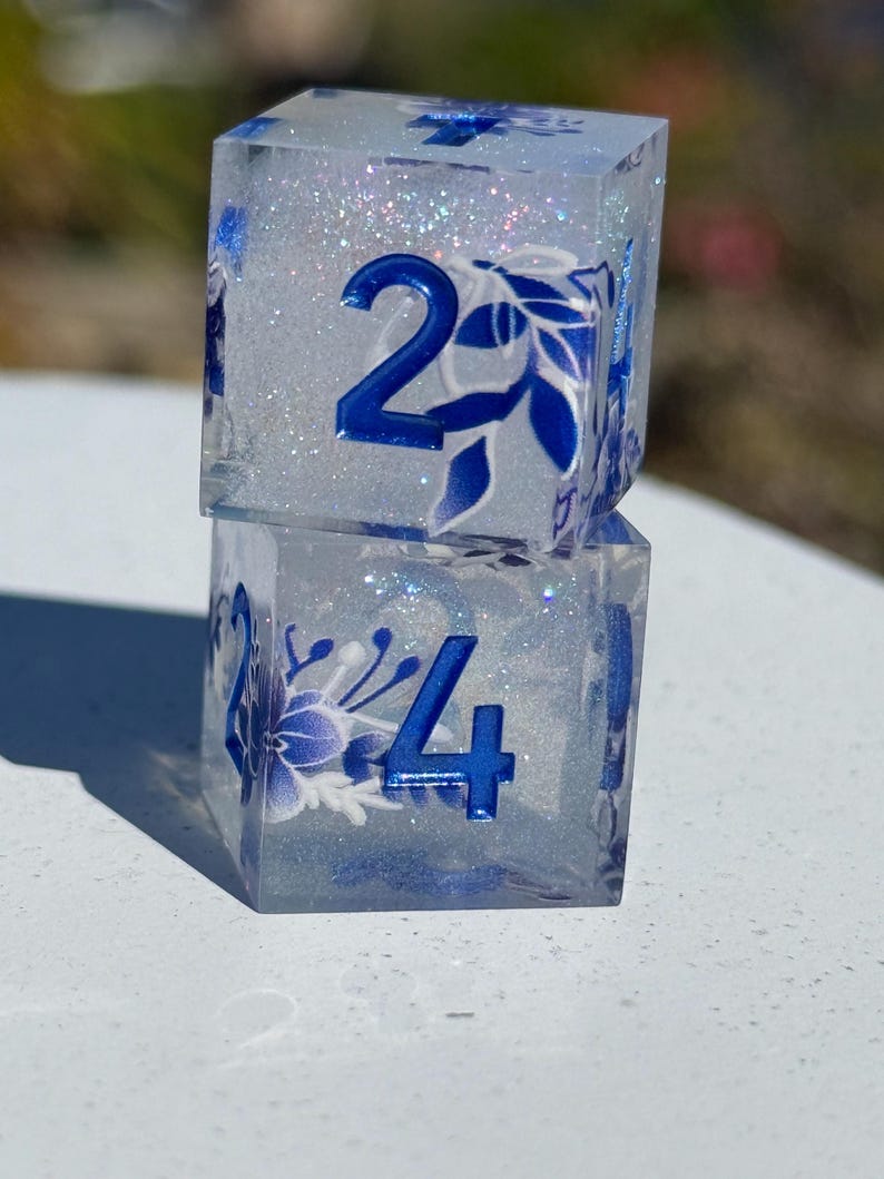 May include: Two clear, square dice stacked on top of each other. The dice have blue numbers and floral designs. The interior of the dice is filled with glitter. The top die shows the number 2, and the bottom die shows the number 4.