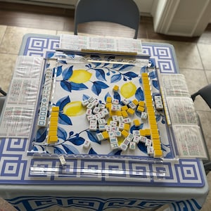 May include: A Mahjong game set on a table with a blue and white geometric pattern tablecloth. The game board features a lemon and leaf design. Yellow and white tiles are scattered across the board, with several stacks of tiles and score cards.
