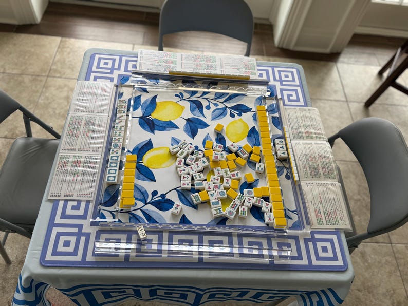 May include: A Mahjong game set on a table with a blue and white geometric pattern tablecloth. The game board features a lemon and leaf design. Yellow and white tiles are scattered across the board, with several stacks of tiles and score cards.